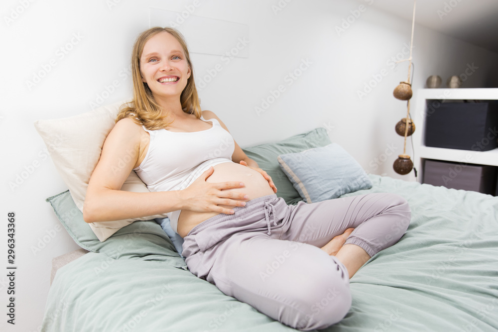 Cheerful expectant mother feeling happy at home. Young pregnant woman relaxing in bed, holding naked belly, looking ta camera, smiling. Pregnancy concept Cheerful expectant mother feeling happy at home. Young pregnant woman relaxing in bed, holding naked belly, looking ta camera, smiling. Pregnancy concept