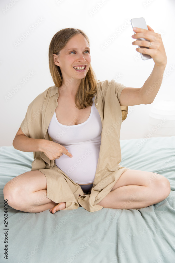 Joyful excited expectant mother showing her big belly while talking through video call. Pregnant woman sitting on bed at home, holding mobile phone, pointing at tummy. Phone call concept Joyful excited expectant mother showing her big belly while talking through video call. Pregnant woman sitting on bed at home, holding mobile phone, pointing at tummy. Phone call concept