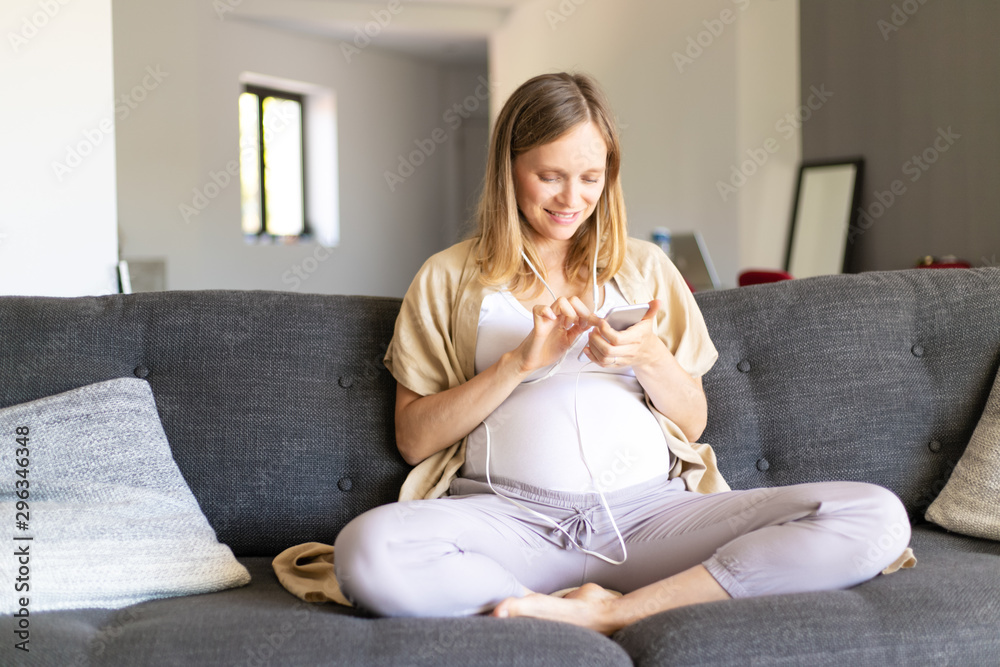 Happy expectant mother watching content on smart phone. Pregnant woman wearing earphones connected with cellphone, sitting on couch, using device, smiling. Communication concept Happy expectant mother watching content on smart phone. Pregnant woman wearing earphones connected with cellphone, sitting on couch, using device, smiling. Communication concept