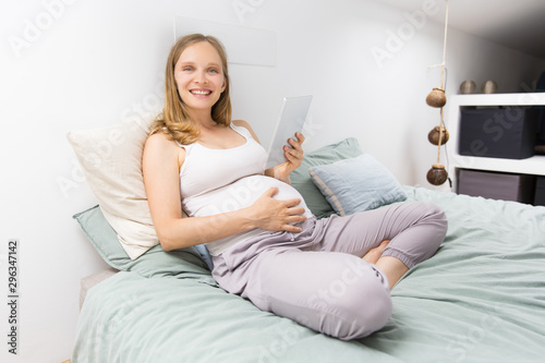 Happy pregnant woman reading book on tablet at home. Young woman resting in bed, holding tablet, touching belly, looking at camera and smiling. Pregnancy or leisure concept