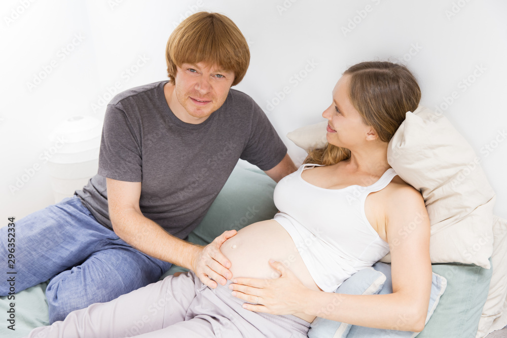 Happy expectant couple enjoying leisure time at home. Young man touching pregnant wife belly and looking at camera. New life concept Happy expectant couple enjoying leisure time at home. Young man touching pregnant wife belly and looking at camera. New life concept