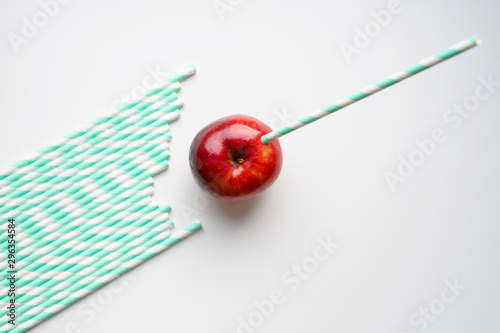 a ripe juicy bright red apple and in it a striped white and green paper drinking straw, and a pile of drinking straws nearby, located on a white background