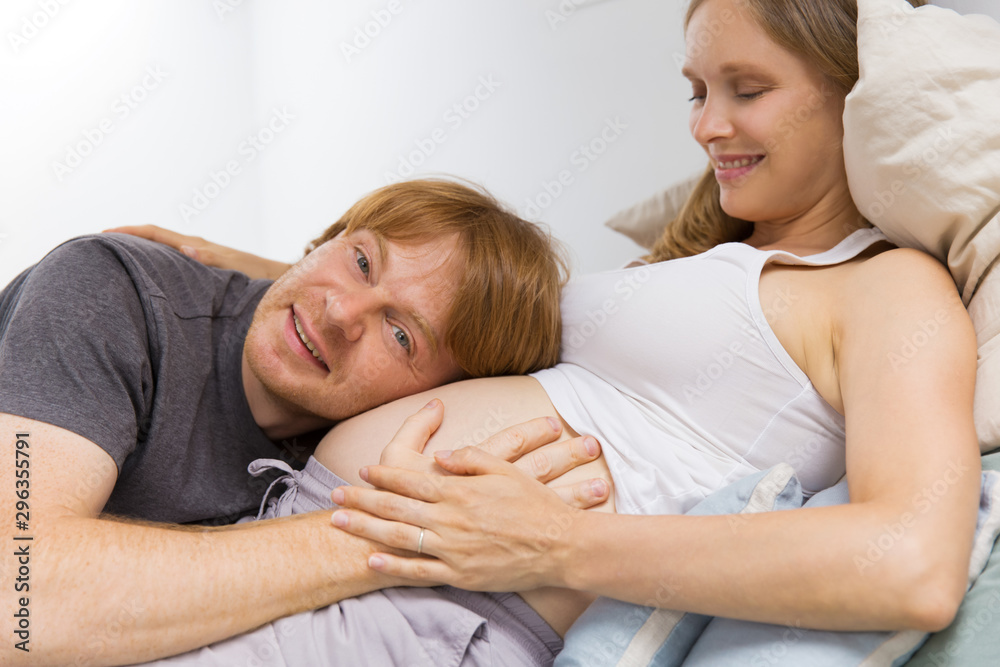 Cute expectant couple enjoying leisure time at home. Happy future dad applying ear to pregnant wife belly, smiling, looking at camera. Family and pregnancy concept Cute expectant couple enjoying leisure time at home. Happy future dad applying ear to pregnant wife belly, smiling, looking at camera. Family and pregnancy concept