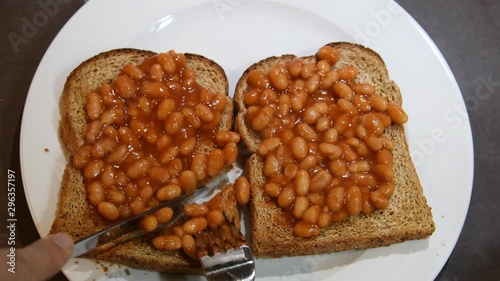 Beans on toast. Hand appearing holding knife and fork and cutting toast with beans on them. Comfort food. Two slices of brown whole meal toast topped with baked beans. Simple British breakfast meal.