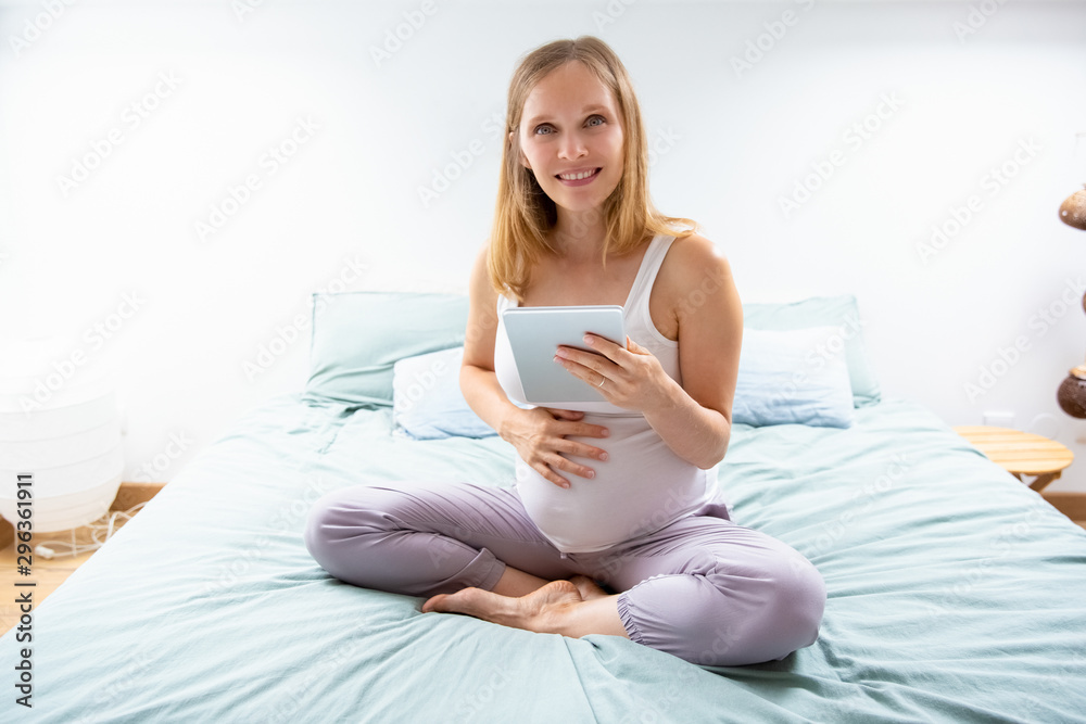 Happy expectant mom with tablet posing in bedroom. Pregnant woman sitting on bed, holding digital device, touching belly, looking at camera. Internet connection at home concept