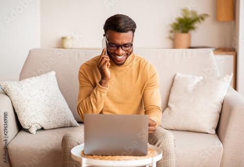 Afro Man Using Laptop Having Pleasant Phone Conversation At Home