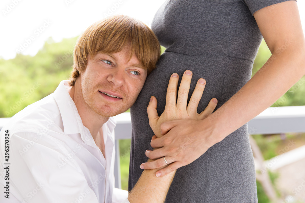 Fototapeta premium Excited focused expectant dad listening to baby bump on terrace. Man applying ear to pregnant wife belly. Pregnant couple concept