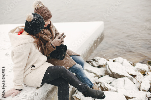 two bright and merry girls sitting in the frozen snowy park neaw water and use the phone photo