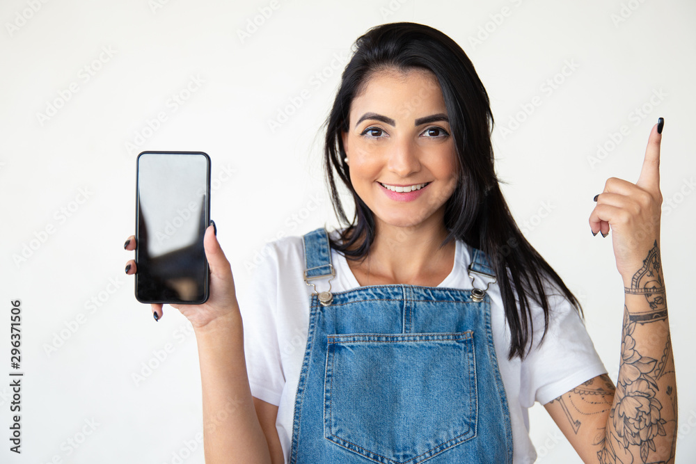 Smiling young woman holding smartphone. Front view of cheerful brunette holding smartphone and pointing up with finger. Technology concept Smiling young woman holding smartphone. Front view of cheerful brunette holding smartphone and pointing up with finger. Technology concept