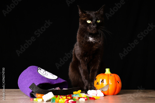 A black cat standing on top of a wooden table next to candy and Halloween decorations