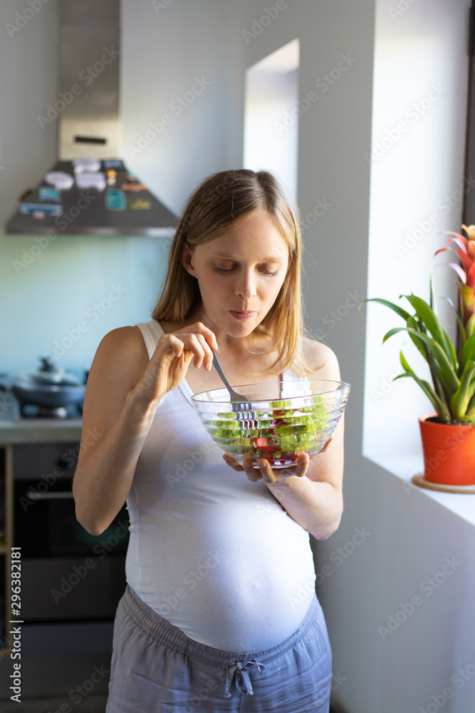 Focused expectant mother enjoying healthy lunch. Pregnant woman standing in kitchen, holding bowl, eating salad. Pregnancy and organic diet concept Focused expectant mother enjoying healthy lunch. Pregnant woman standing in kitchen, holding bowl, eating salad. Pregnancy and organic diet concept