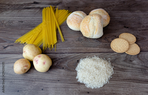 Common bad carbohydrates to avoid such as white bread, pasta, rice, potatoes and buscuits laying on top of a wooden table viewed from above