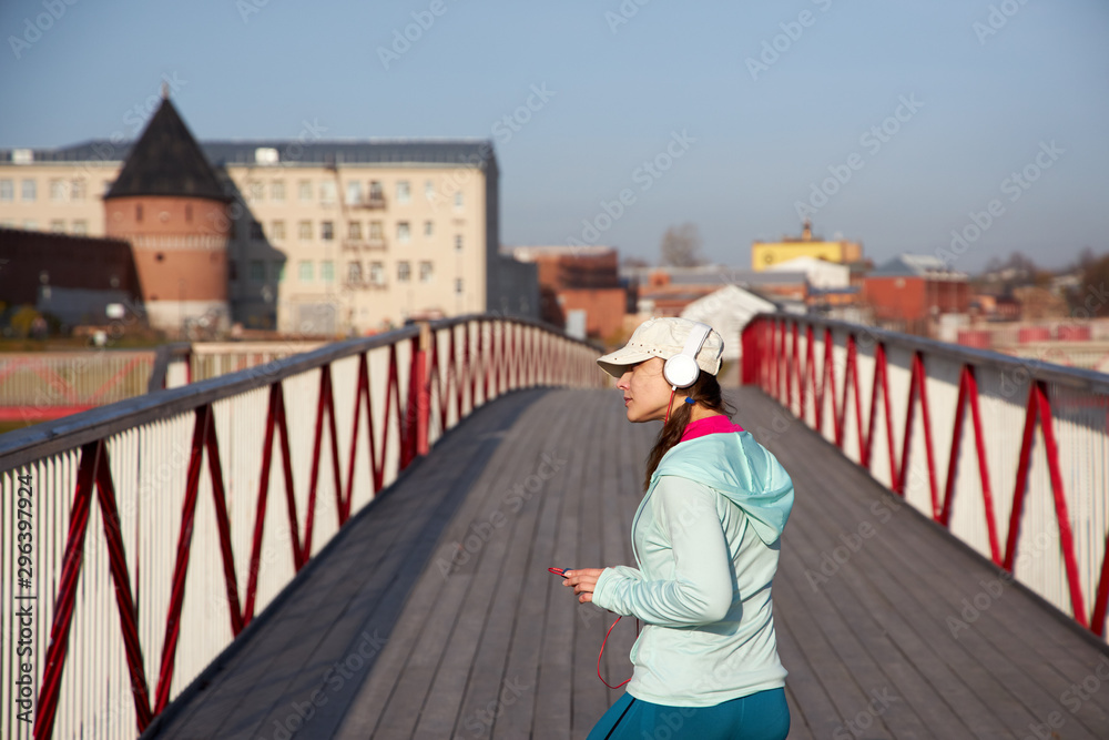 Woman relaxing on a wooden bridge after cardio exercise outdoors, taking care of her body, staying healthy and fit, in a white baseball hat, motion in headphones side view listening to music