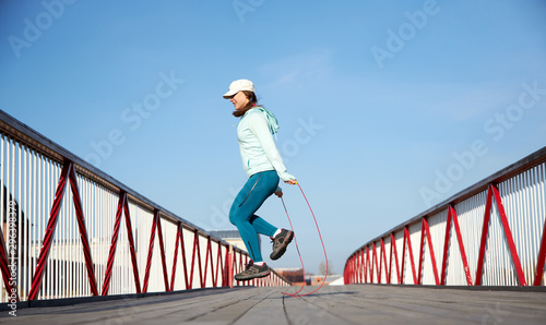 woman jumping rope on a wooden bridge, cardio exercise outdoors, taking care of her body, staying healthy and fit, in a white baseball hat, motion stamina health 
