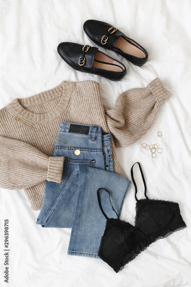 Blue jeans, beige knitted sweater, black lace bra, black loafers or flat  shoes lying on bed