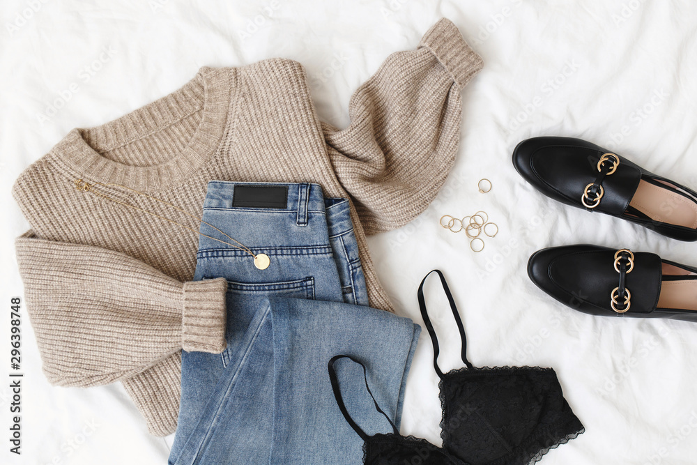 Blue jeans, beige knitted sweater, black lace bra, black loafers or flat  shoes lying on bed