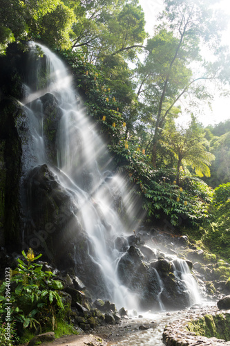 Waterfall with bright sun rays bursting through the mist created by the falling water at the Parque Natural da Ribeira dos Caldeirões, São Miguel island, Azores. Daytime vertical long exposure image. © RichHiggins