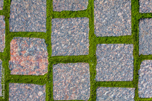old gray red granite stone pavement made of square stone bricks with sprouted green moss. rough surface texture