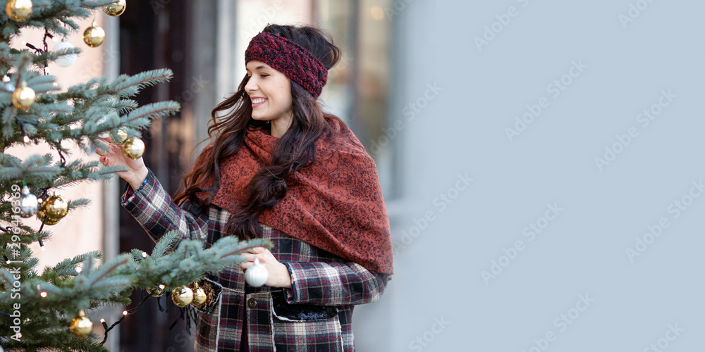 Beautiful joyful woman portrait in a city. Smiling  girl wearing warm clothes and hat  in winter. Christmas time with natural decorative fir tree. Copy space - obrazy, fototapety, plakaty 