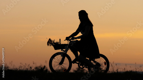 Young woman silhouette arrange flowers bouquet on bike basket, girl ride, sunset