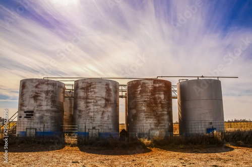 Old rusty holding tanks with shadows at oil well location on flat farm land under dramatic sky at golden hour