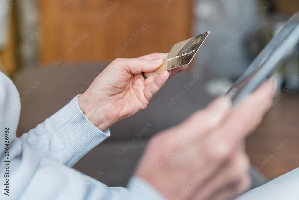 Cropped of senior woman holding bank card while shopping online on digital tablet sitting on sofa at home