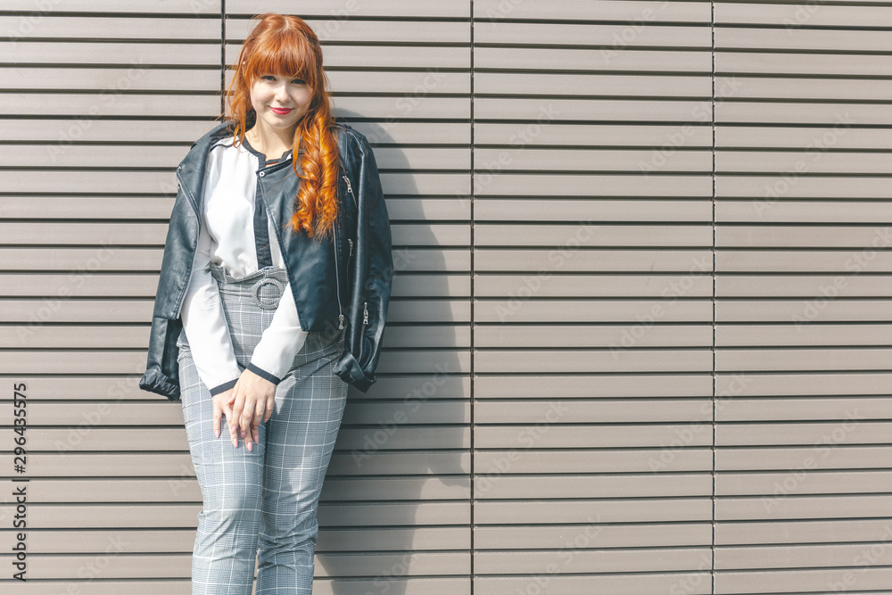 Cute young girl in a black leather jacket with curly red hair and a fringe stands near the modern wall on a sunny day and looks at the camera Cute young girl in a black leather jacket with curly red hair and a fringe stands near the modern wall on a sunny day and looks at the camera