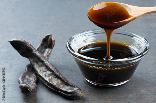carob molasses in glass bowl and in wooden spoon and carob pods on rustic background, locust bean healthy food, Ceratonia siliqua ( harnup ) photo