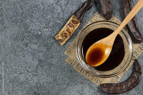 Foto carob molasses in glass bowl and in wooden spoon and carob pods on rustic backgr