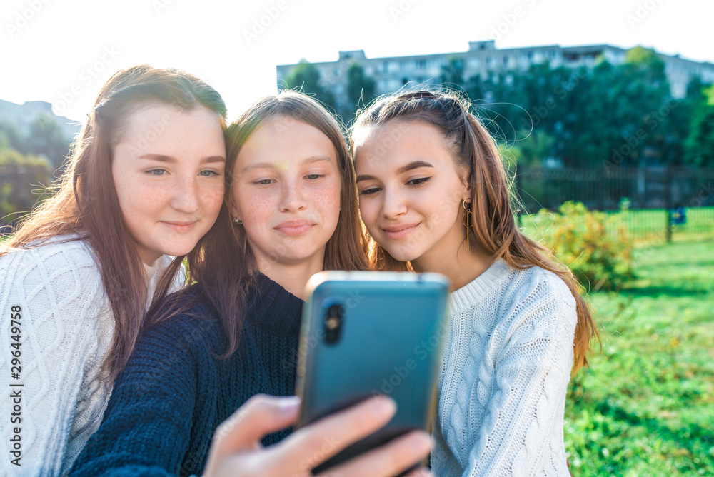 Three girls schoolgirls girlfriends, watch videos stories social networks, photograph camera phone. Video recording video call online application on Internet summer city nature. Autumn warm sweaters. Three girls schoolgirls girlfriends, watch videos stories social networks, photograph camera phone. Video recording video call online application on Internet summer city nature. Autumn warm sweaters.