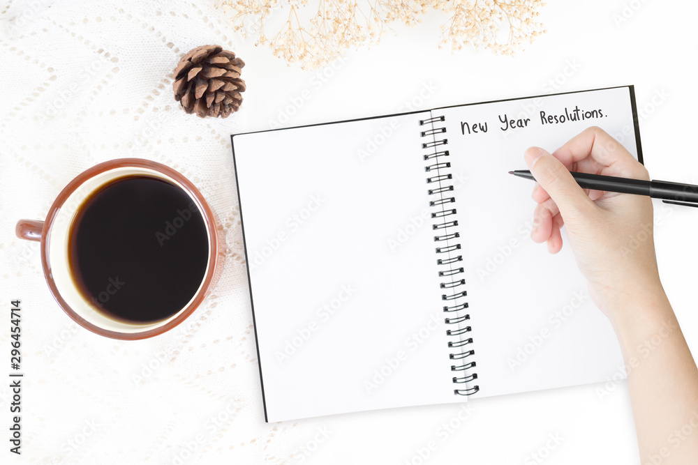 Top view of cup of coffee and pine tree and dry flower over white table cover and the right hand is writing down the text for new year goals and resolutions list on white paper note. Top view of cup of coffee and pine tree and dry flower over white table cover and the right hand is writing down the text for new year goals and resolutions list on white paper note.