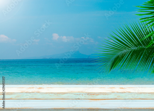 Fototapeta Naklejka Na Ścianę i Meble -  Vintage summer plank blue grunge board table with clear blue sea with sunny sky with cloud and green coconut tree leaves in  background.