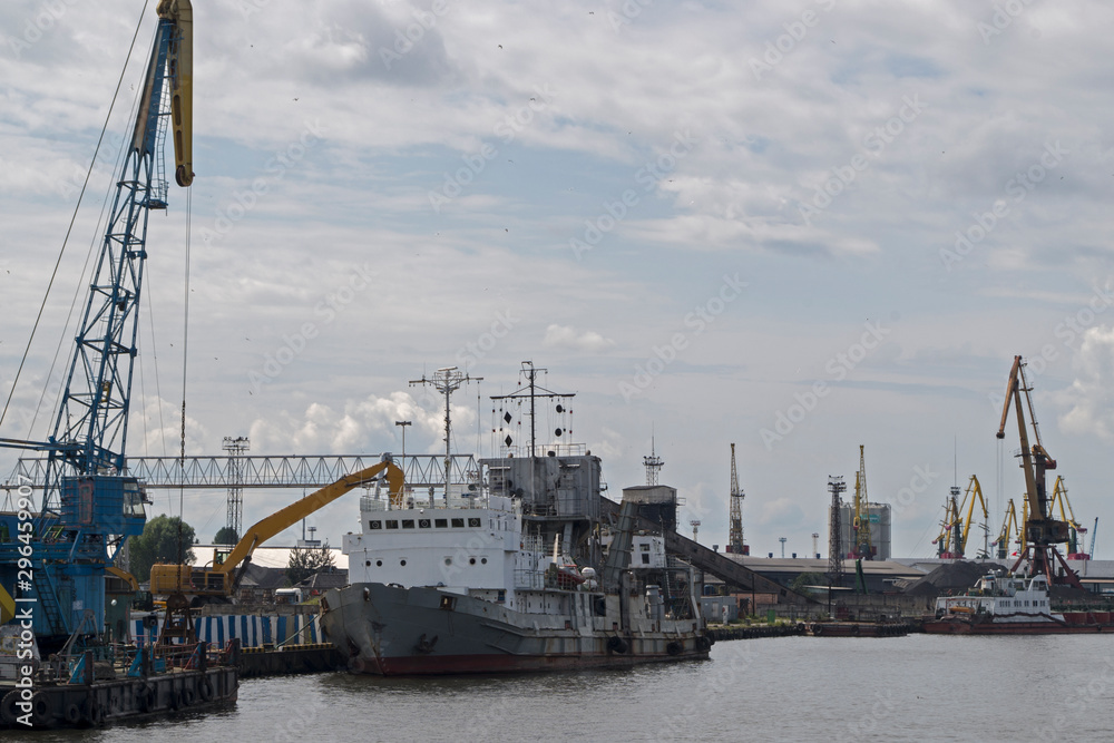 Coal loading or unloading of a grey bulk carrier with a white deck using an excavator in the port, nearby loading platforms and port loading cranes can be seen on the background a cloudy blue sky