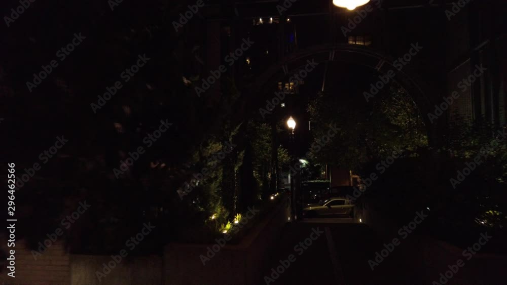 Moving down dark pathway lit by hanging lamps and out to the street with man walking by and cars parked. Moving down dark pathway lit by hanging lamps and out to the street with man walking by and cars parked.