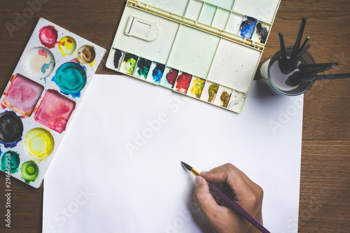 Man hand holding paint brush on white paper with colorful watercolor and equipment on wooden background.