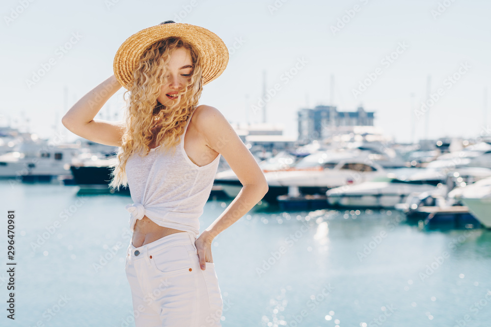 Charming young girl with long blond curly hair in a white blouse and shorts with a straw hat and dances against the background of the ocean
