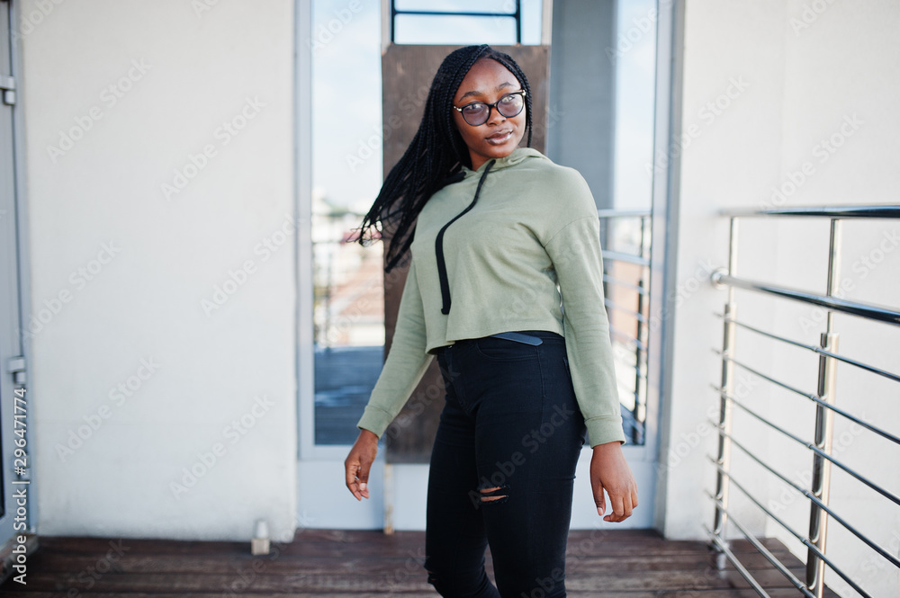 City portrait of positive young dark skinned female wearing green hoody and eyeglasses standing at balkony. City portrait of positive young dark skinned female wearing green hoody and eyeglasses standing at balkony.