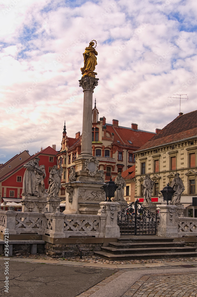 Beautiful landscape view of Plague Column against cloudy sky. Ancient colorful building at the background. The Rotovz Town Hall Square in Maribor, Lower Styria, Slovenia