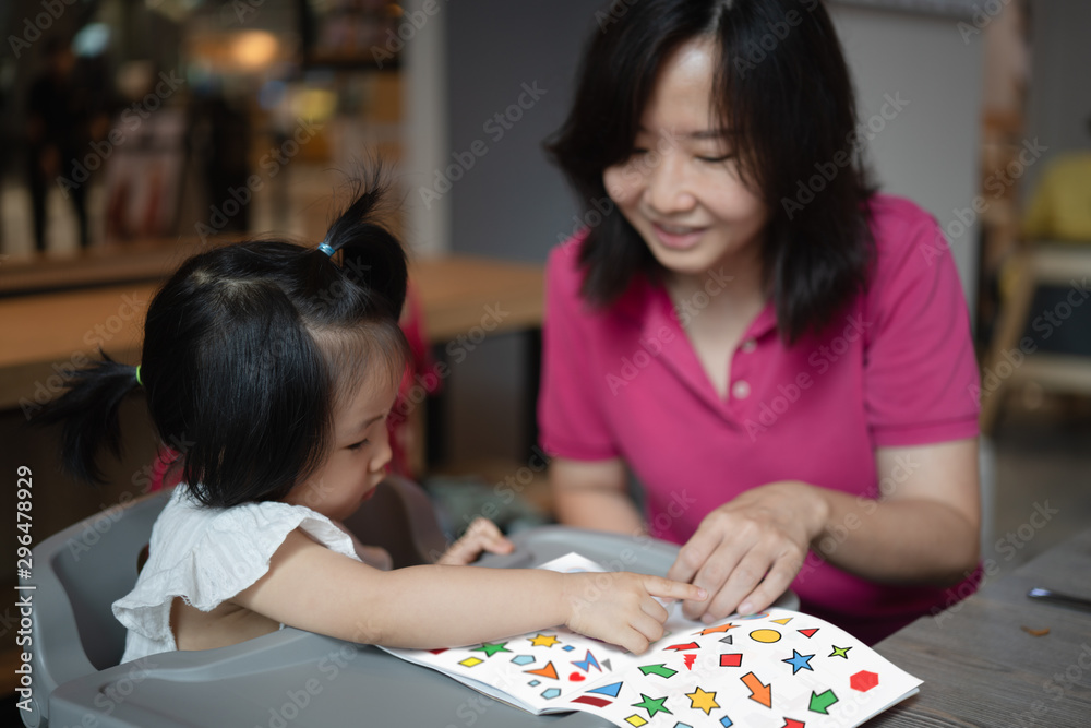 Fototapeta premium concept of Asian mother and 2 years old spending time together learning and doing activities, avoiding screen time by using paper book and sticker, while the daughter concentrate and enjoy learning.