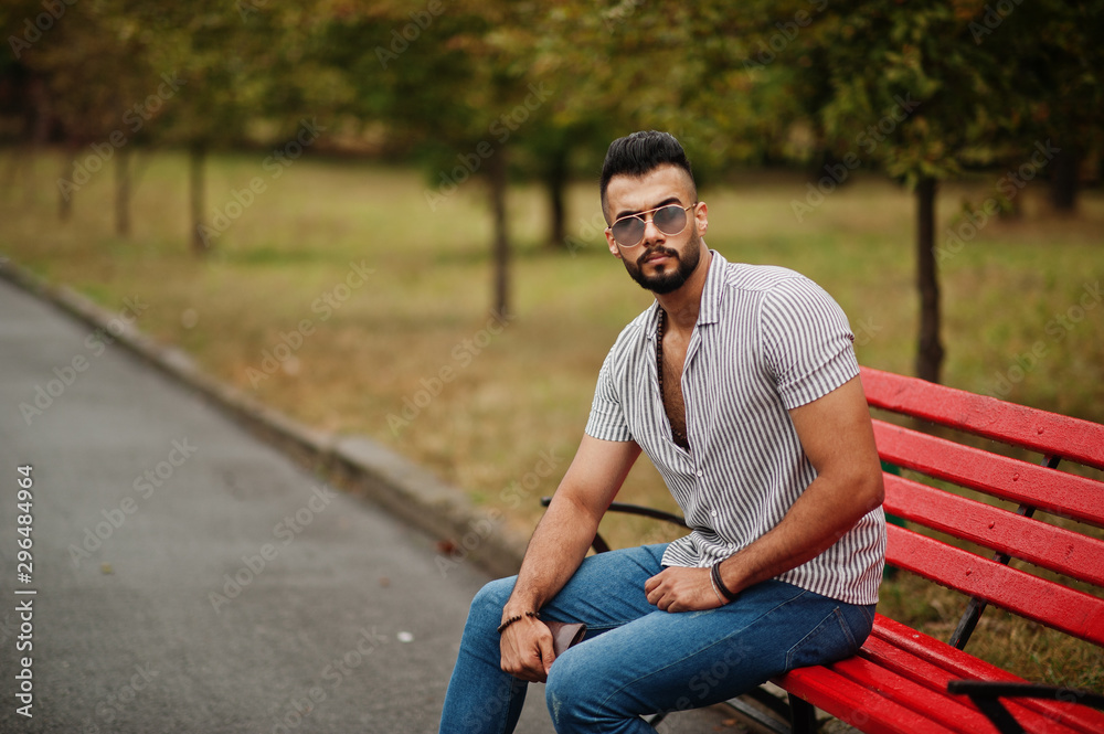 Fashionable tall arab beard man wear on shirt, jeans and sunglasses sitting on red bench at park. Fashionable tall arab beard man wear on shirt, jeans and sunglasses sitting on red bench at park.
