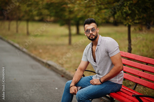Fashionable tall arab beard man wear on shirt, jeans and sunglasses sitting on red bench at park. © AS Photo Family