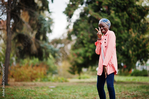 Young modern fashionable, attractive, tall and slim african muslim woman in hijab or turban head scarf and pink coat posed at park.