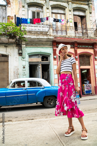Stylish woman in a bright dress and white hat next to retro car on a city street of Cuba