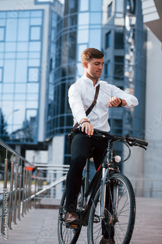 Looks at watch. Businessman in formal clothes with black bicycle is in the city