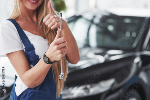 Shows thumb up and holds wrench in other hand. Nice blonde woman repairer is on her work. Indoors at car shop