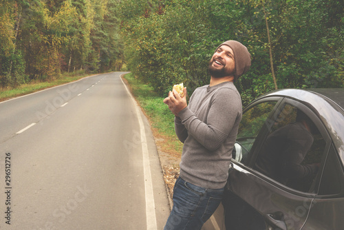Young guy eating a burger near a car on an empty road. Food on the trip. Food on the go. Autumn travel. Fast food.