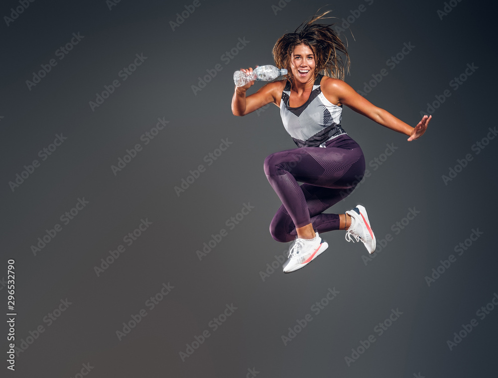 Exited cheerful woman is jumping at studio while holding the bottle of water over grey background.