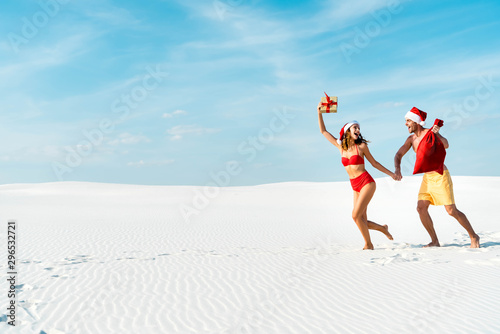 sexy and smiling girlfriend holding gift and boyfriend with santa sack on beach in Maldives