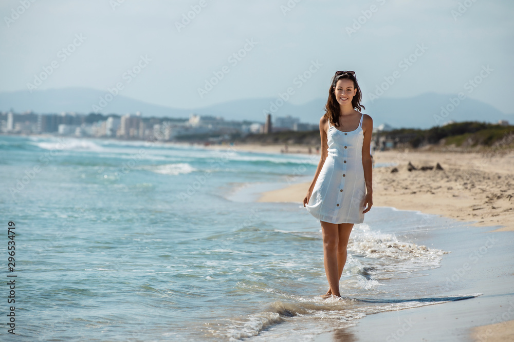 young brunette woman wearing white dress and sunglasses, walking on a beach at the mediterranean sea, or Caribbean 
