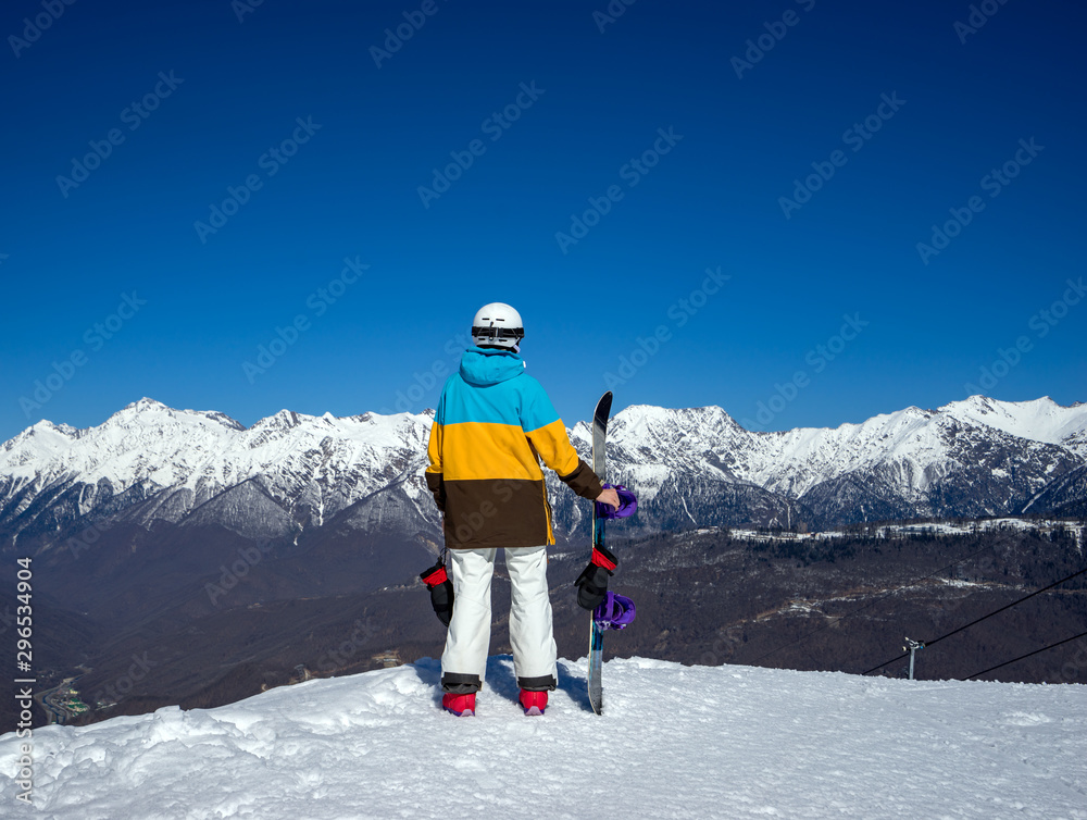 Woman snowboarder standing at the edge of the slope and admiring mountain scenery of the Caucasus mountains in Krasnaya Polyana